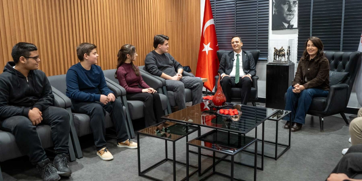 Modern bir ofis ortamında, ahşap çıtalı bir duvarın önünde yer alan gri koltuklarda oturan dört genç, Bornova Belediye Başkanı Mustafa İduğ ve bir kadın yetkili ile sohbet ediyor. Odanın köşesinde Türk bayrağı, duvarda Mustafa Kemal Atatürk’ün portresi ve bir kaide üzerinde küçük bir heykel bulunuyor. Ortadaki cam sehpanın üzerinde kırmızı dekoratif bir obje ve çiçekler yer alıyor.