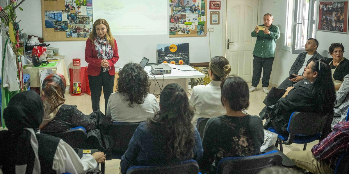 Kırmızı ceketli bir kadın, bir sınıfta veya toplantı odasında, sıfır atık konulu bir sunum yaparken katılımcılara hitap ediyor. Katılımcılar sandalyelerde oturarak kadını dinliyor, odanın köşesinde bir kişi ise cep telefonuyla fotoğraf çekiyor. Arka planda üzerinde çeşitli fotoğrafların bulunduğu bir pano, projeksiyon perdesi ve 