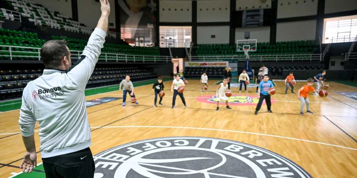 Bornova Belediyesi logolu eşofman üstü giyen bir antrenör, kapalı bir spor salonunda basketbol topu süren bir grup çocuğa eğitim veriyor. Çocuklar parke zemin üzerinde dizilmiş durumda ve her birinin elinde bir basketbol topu bulunuyor. Arka planda yeşil koltuklu tribünler ve salonun ortasında büyük bir logo yer alıyor.