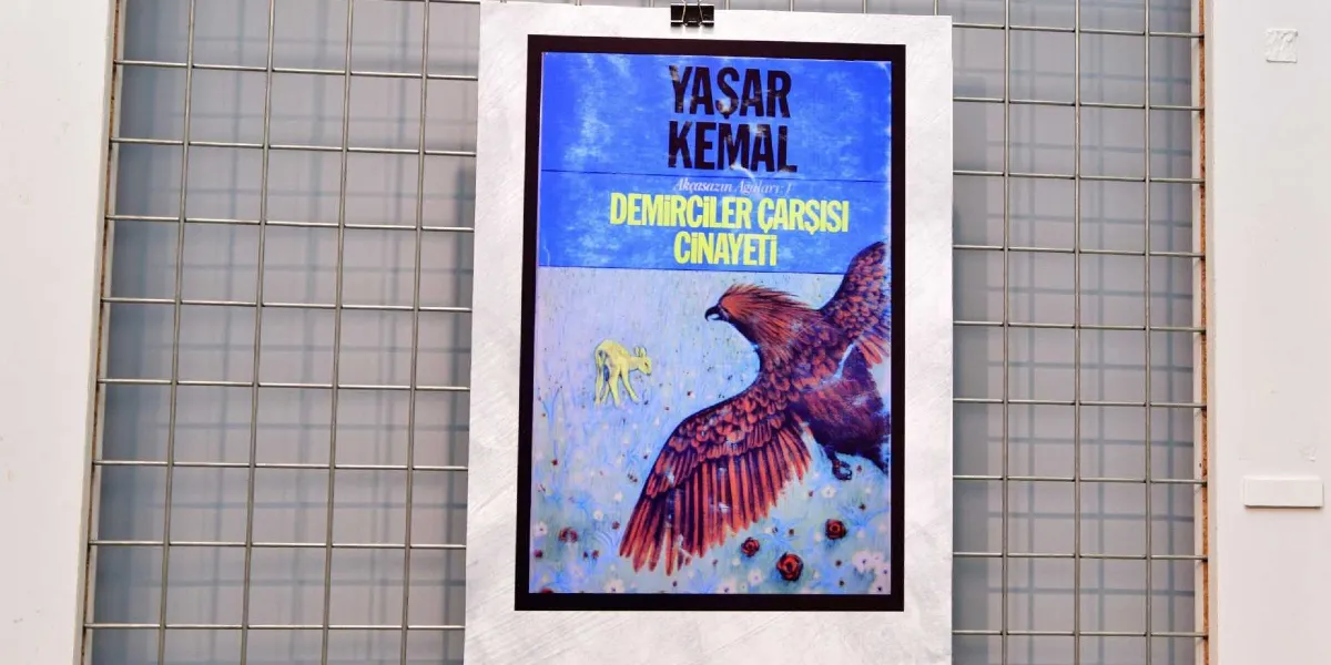 Metal bir ızgara pano üzerine asılmış, Yaşar Kemal'in Akçasazın Ağaları serisinin ilk kitabı olan Demirciler Çarşısı Cinayeti romanının kapak görseli. Kapakta, mavi bir gökyüzü altında kanatlarını açmış büyük bir yırtıcı kuş ve yanında küçük, sarı bir kuzu figürü yer alıyor.