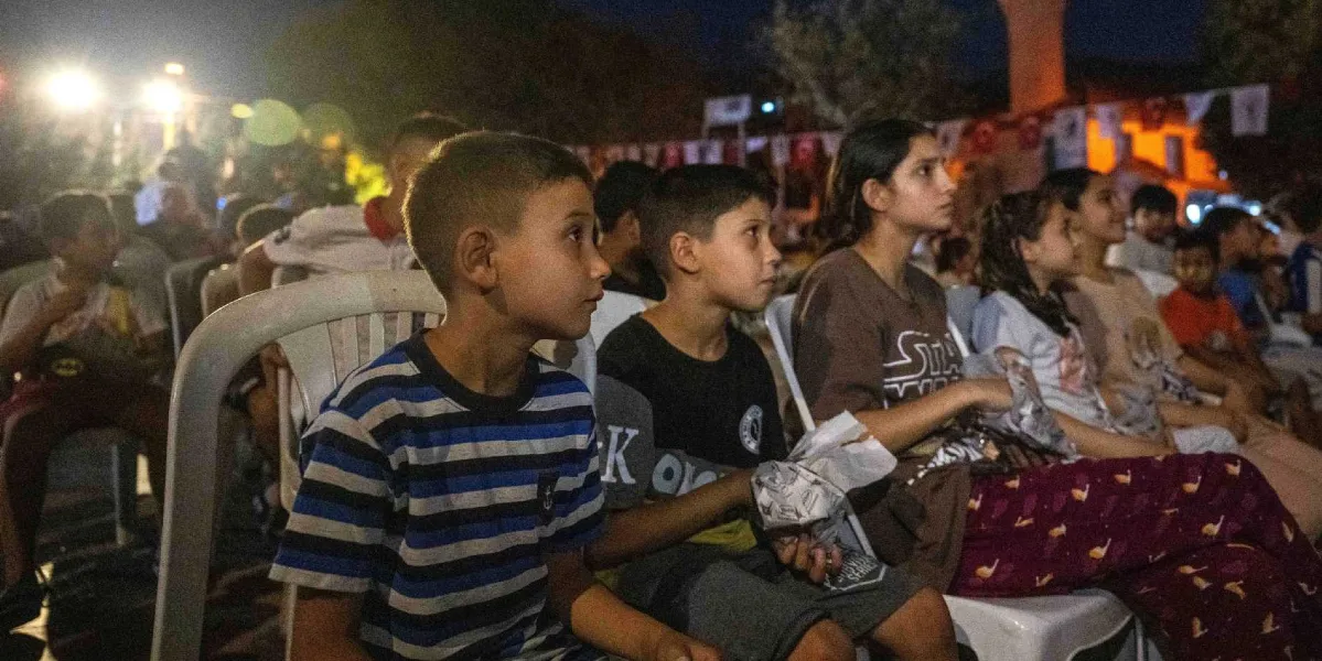 Açık havada düzenlenen bir etkinlikte beyaz plastik sandalyelerde yan yana oturmuş, dikkatle karşıya bakan çocuklar ve gençler. Ön planda mavi-beyaz çizgili tişörtlü bir erkek çocuğu, yanında siyah tişörtlü bir başka çocuk ve ellerinde patlamış mısır paketleri tutan genç kızlar yer alıyor. Arka planda ise akşam karanlığında ışıklandırılmış bir alan ve asılı bayraklar görülüyor.