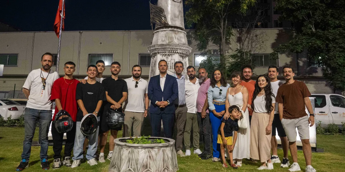 Gece vakti, Bornova'daki Abide-i Hürriyet anıtının önünde bir araya gelmiş, farklı yaş gruplarından oluşan kalabalık bir grup insan yan yana durarak poz veriyor. Gruptakilerin çoğu günlük kıyafetler giymiş, bazıları ellerinde motosiklet kaskları tutuyor. Arka planda aydınlatılmış anıtın gövdesi ve ağaçlar görülürken, sağ tarafta beyaz bir minibüs park halinde bekliyor.