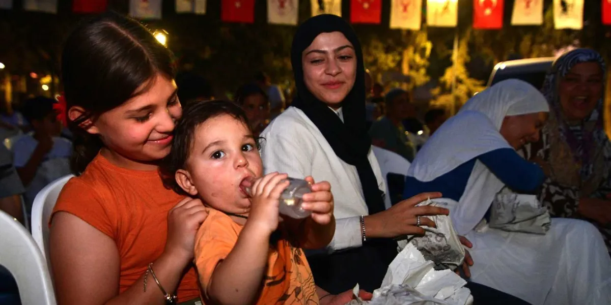 Açık havada düzenlenen bir etkinlikte, yan yana oturan ve gülümseyen bir grup insan. Ön planda turuncu tişörtlü bir kız çocuğu, kucağındaki küçük bir bebeğe biberonla su içiriyor. Yanlarında başörtülü bir kadın ve arkalarında neşeyle gülen başka izleyiciler bulunuyor. Arka planda ise gece karanlığında asılı duran bayraklar ve ışıklar seçiliyor.
