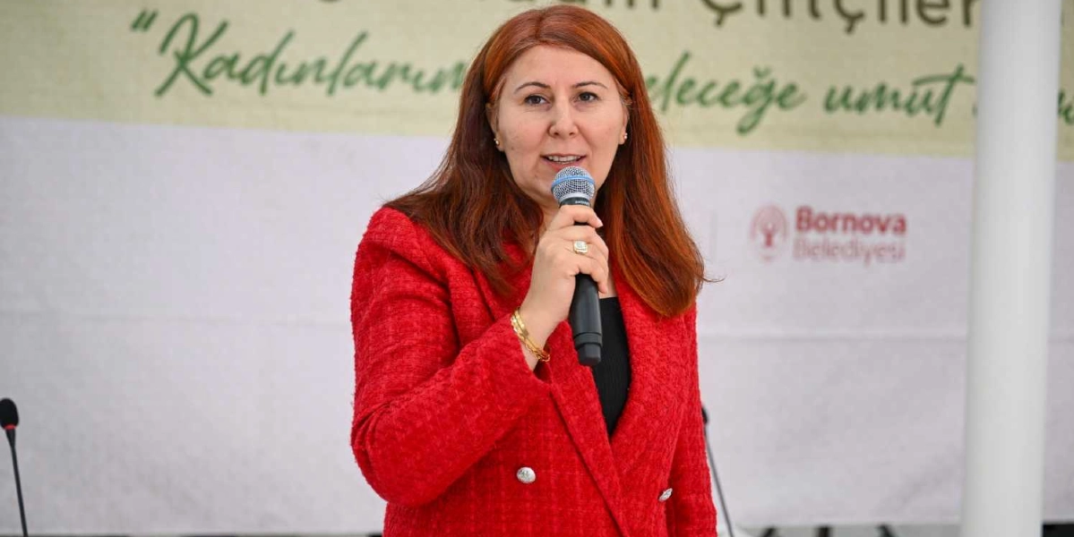 Kızıl saçlı, kırmızı ceketli bir kadın, elinde mikrofonla konuşma yapıyor. Arka planda Bornova Belediyesi logosunun ve üzerinde 