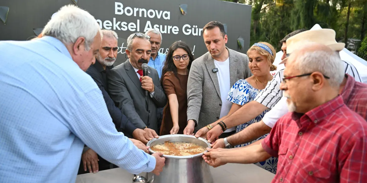 Bornova Geleneksel Aşure Günü etkinliğinde, bir grup insan ellerini büyük bir aşure kazanının üzerine koymuş şekilde duruyor. Gruptan biri mikrofonla konuşma yaparken, diğerleri aşure kazanını çevreleyerek birlik mesajı veriyor. Arka planda üzerinde Bornova Geleneksel Aşure Günü yazılı bir pano yer alıyor.