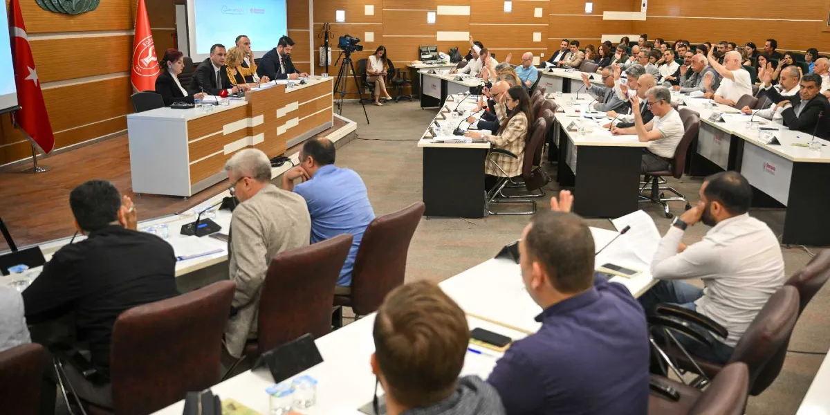 Bornova Belediyesi meclis salonunda düzenlenen toplantıda, katılımcıların birçoğu ellerini kaldırarak oylamaya katılıyor. Salonun ön kısmındaki masada yetkililer otururken, diğer katılımcılar U düzenindeki sıralarda yer alıyor. Arka planda Türk bayrağı ve Bornova Belediyesi logosu bulunan bir flama yer alıyor.