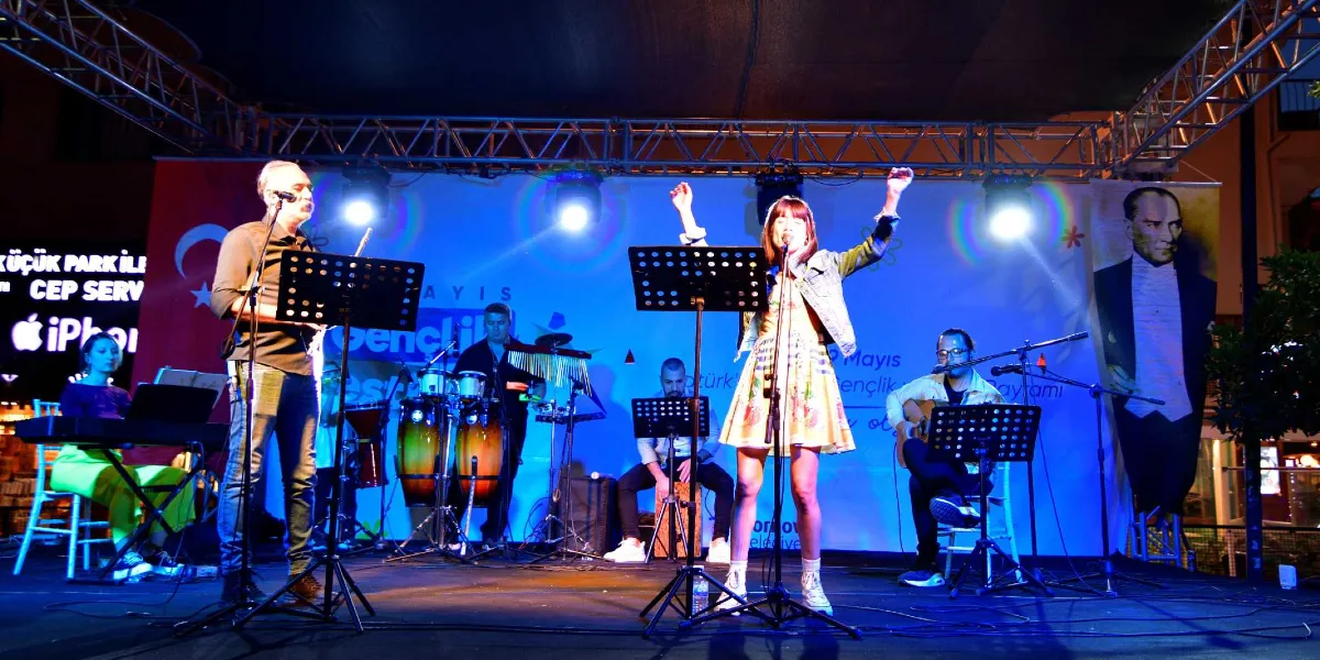 Bornova'da düzenlenen 19 Mayıs Atatürk'ü Anma, Gençlik ve Spor Bayramı kutlamaları kapsamında, açık hava sahnesinde performans sergileyen bir müzik grubu. Sahne üzerinde soldan sağa doğru klavye çalan bir kadın, mikrofon başında şarkı söyleyen bir erkek, perküsyon çalan bir müzisyen, kucağında kutu davul (cajon) ile oturan bir başka müzisyen, ellerini havaya kaldırarak şarkı söyleyen bir kadın solist ve gitar çalan bir erkek müzisyen yer alıyor. Arka planda üzerinde Atatürk portresi ve 