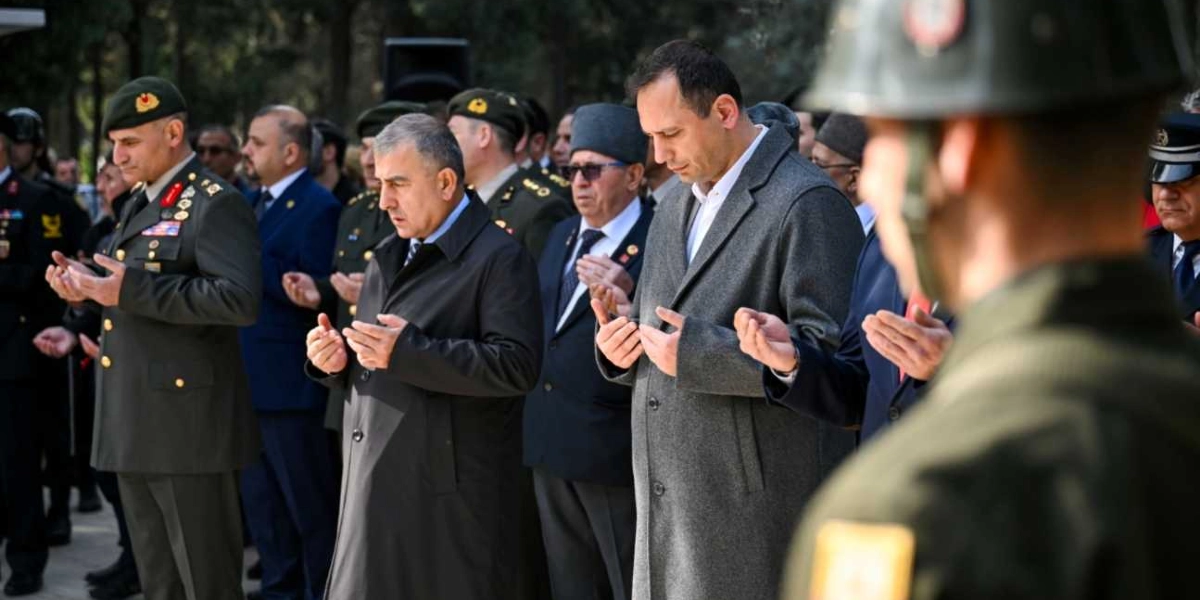 Resmi üniformalı askerler, takım elbiseli sivil yetkililer ve vatandaşların bir arada bulunduğu bir anma töreninde, katılımcılar ellerini açmış dua ediyor. Ön planda gri paltolu bir erkek, yanında takım elbiseli ve kalpaklı bir kişi ile üniformalı bir subay yer alıyor. Arka planda ise törene katılan diğer kişiler ve askeri personel görülüyor.