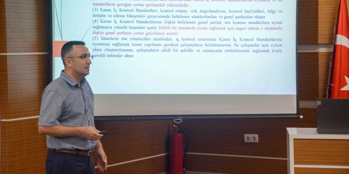 Gözlüklü, kısa saçlı ve gri kısa kollu gömlek giymiş bir erkek, projeksiyon perdesinin önünde sunum yapmaktadır. Perdede 