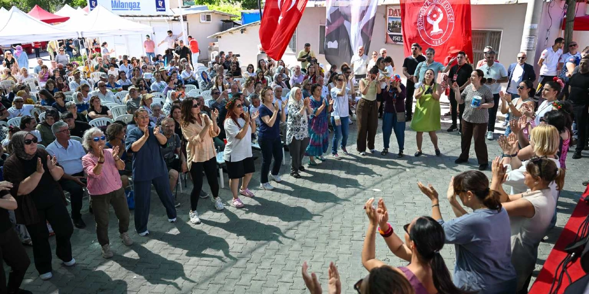 Açık havada düzenlenen bir etkinlikte, Bornova Belediyesi bayrağının altında toplanan kalabalık bir grup insan, alkışlarla eğleniyor. Parke taşlı bir meydanda, beyaz plastik sandalyelerde oturan izleyicilerin önünde, ayakta duran kadınlar ve erkekler neşeyle ellerini çırpıyor. Arka planda beyaz çadırlar, Türk bayrakları ve çeşitli katılımcılar görülüyor.