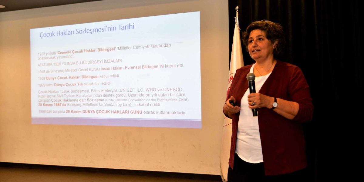Kısa saçlı bir kadın, elinde mikrofonla bir sunum yapmaktadır. Arkasındaki projeksiyon perdesinde 
