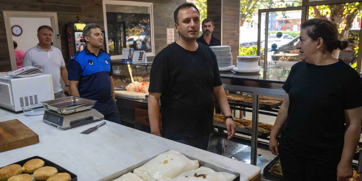 Belediye başkanı, yanında bir zabıta memuru ve diğer görevlilerle birlikte bir fırın veya unlu mamuller dükkanında denetim yaparken, tezgahın arkasındaki bir kadın çalışanla konuşuyor. Tezgahın üzerinde pişmeye hazır hamur işleri ve bir tartı bulunuyor.