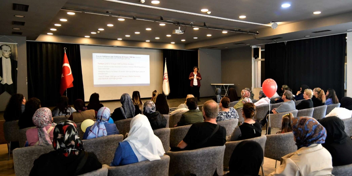 Kapalı bir konferans salonunda, sahnedeki bir konuşmacıyı dinleyen kalabalık bir izleyici grubu. Sahnedeki projeksiyon perdesinde Çocuk Haklarına Dair Sözleşme ile ilgili bilgiler yer alıyor. Salonun bir köşesinde Türk bayrağı, diğer köşesinde ise Bornova Belediyesi'ne ait olduğu anlaşılan bir flama bulunuyor. İzleyicilerin arasında elinde kırmızı bir balon tutan bir kişi dikkat çekiyor. Salonun sol duvarında ise Atatürk portresi asılı.