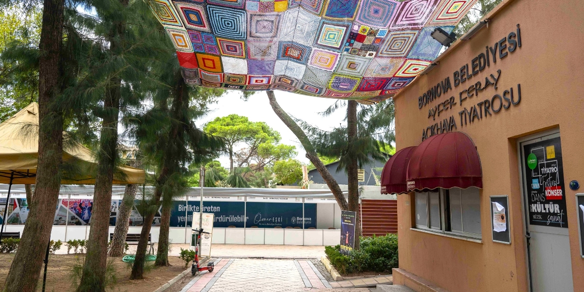 Bornova Belediyesi Ayfer Feray Açıkhava Tiyatrosu'nun girişinde, ağaçların arasına gerilmiş rengarenk örgü motiflerinden oluşan büyük bir gölgelik bulunuyor. Sağ taraftaki binanın duvarında kurumun ismi yazılı ve giriş kapısında tiyatro, konser, dans, kültür ve sanat etkinliklerini duyuran bir afiş yer alıyor. Arka planda ise üzerinde 