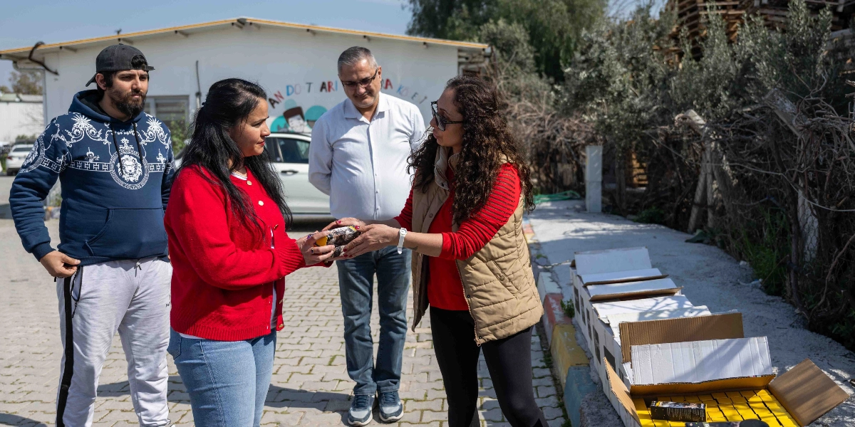 Açık havada, bir barınak veya merkez binasının önünde duran üç kişi, yardım malzemesi olarak görünen küçük paketleri birbirine teslim ediyor. Kırmızı hırkalı bir kadın, elindeki paketi diğer kadına uzatırken, yanlarında duran bir erkek onları izliyor. Sağ tarafta, yerdeki karton kutuların içinde çok sayıda paketlenmiş ürün bulunuyor. Arka planda 