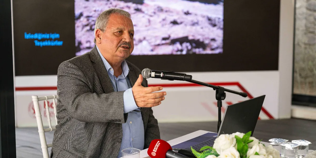 Gri ceketli ve açık mavi gömlekli, bıyıklı orta yaşlı bir erkek, bir masa başında oturarak mikrofon aracılığıyla konuşma yapıyor. Önünde açık bir dizüstü bilgisayar, Bornova Belediyesi logolu bir mikrofon ve beyaz çiçeklerden oluşan bir aranjman bulunuyor. Arka planda ise üzerinde 