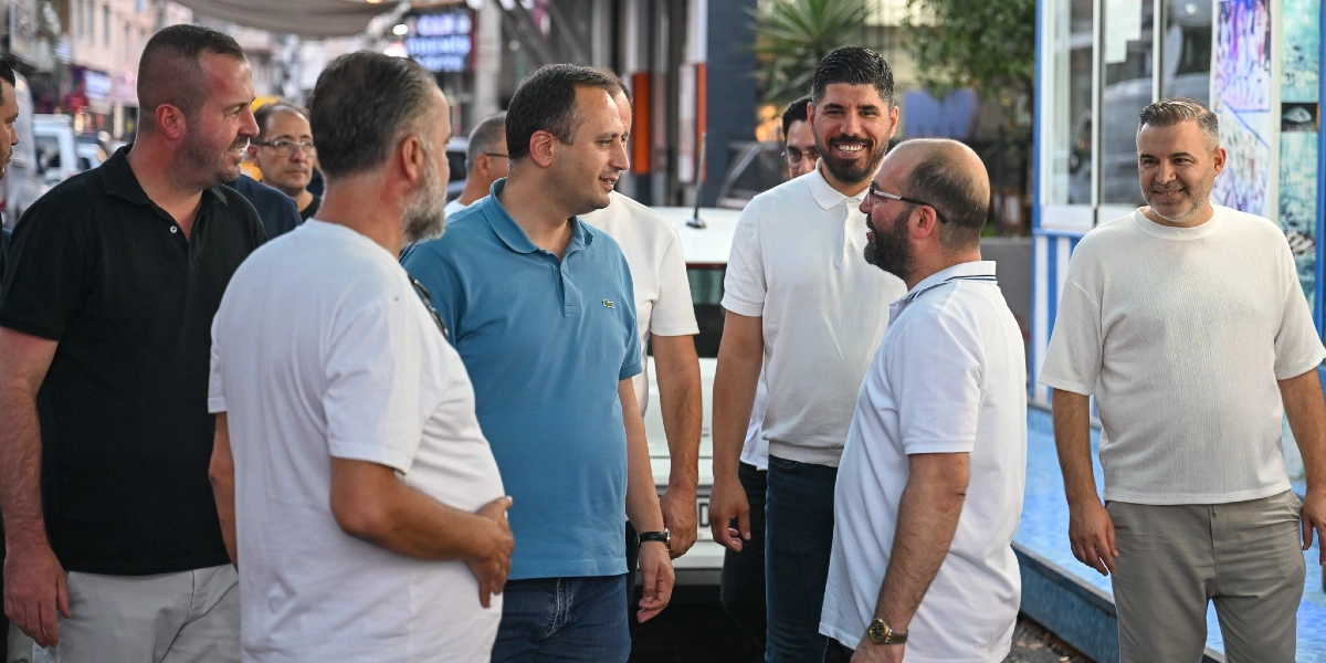 Mavi polo yaka tişört giyen Başkan Eşki, Çamdibi'nde bir sokak üzerinde esnaf ve vatandaşlarla bir araya gelerek sohbet ediyor. Çevresindeki kişilerle samimi bir diyalog içerisinde olan Başkan Eşki'ye, beyaz tişörtlü ve günlük kıyafetli bir grup eşlik ediyor.