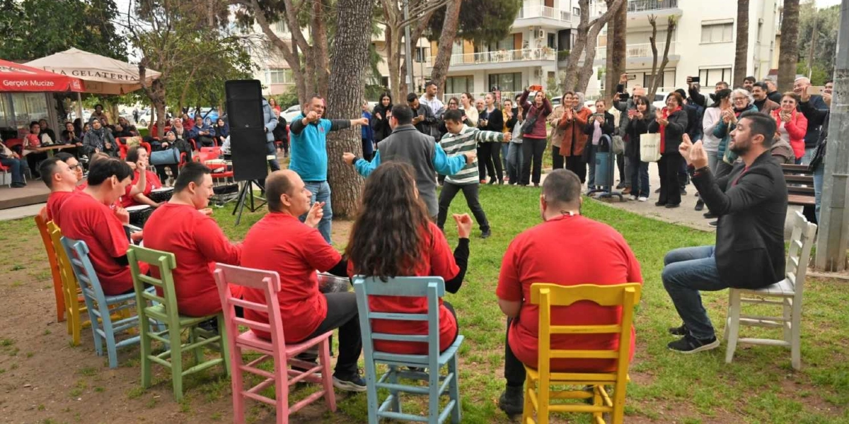 Açık havada, çimenlik bir alanda renkli sandalyelere oturmuş kırmızı tişörtlü bir grup genç, bir eğitmen eşliğinde elleriyle ritim tutuyor. Grubun önünde ayakta duran iki kişi dans ederken, arkada onları izleyen ve alkışlayan kalabalık bir grup yer alıyor. Arka planda ağaçlar, binalar ve bir kafe alanı görülüyor.