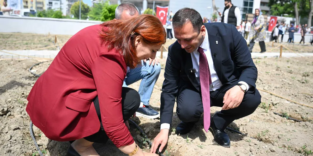Kızıl saçlı bir kadın ve koyu renk takım elbiseli, bordo kravatlı bir erkek, açık havada toprak bir alana çömelmiş şekilde birlikte fidan dikiyor. Arka planda, ellerinde Türk bayrakları taşıyan ve etkinliği izleyen bir grup insan ile ağaçlar görünüyor.