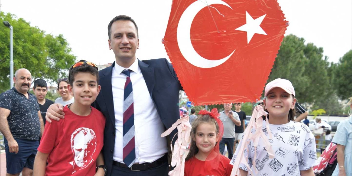 Açık havada düzenlenen bir etkinlikte, üzerinde Türk bayrağı bulunan altıgen bir uçurtmayı tutan bir erkek ve yanlarında duran üç çocuk objektife gülümseyerek bakıyor. Takım elbiseli adamın solunda Atatürk baskılı kırmızı tişört giyen bir erkek çocuk, sağında ise kırmızı kurdeleli bir kız çocuğu ve beyaz desenli tişörtlü başka bir kız çocuğu yer alıyor. Arka planda ise etkinliğe katılan diğer insanlar ve ağaçlar görünüyor.