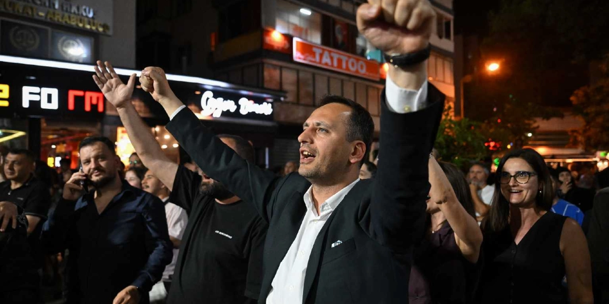 Gece vakti kalabalık bir caddede, siyah ceketli ve beyaz gömlekli bir adamın heyecanla kollarını havaya kaldırarak kutlama yaptığı an. Çevresinde gülümseyen ve destek veren insanlar bulunurken, arka planda ışıklı dükkan tabelaları ve şehir yaşamından kesitler yer alıyor.