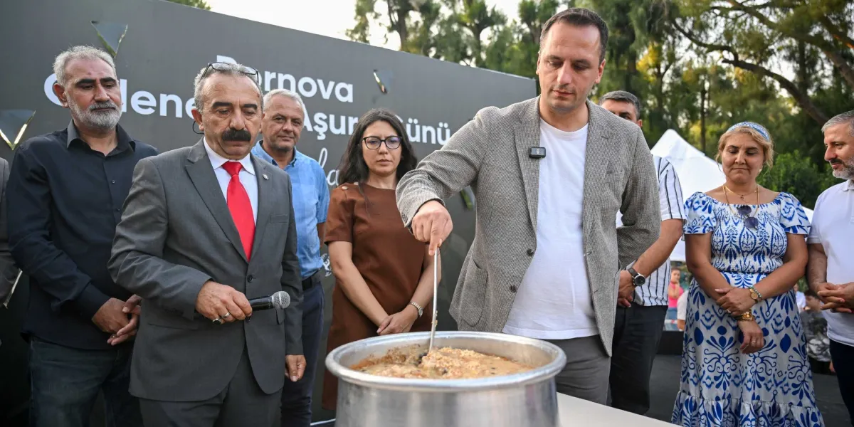 Bornova Belediyesi'nin düzenlediği aşure günü etkinliğinde, gri ceketli bir yetkili büyük bir kazanda aşure karıştırırken, yanında mikrofon tutan bir beyefendi ve diğer katılımcılar izliyor. Arka planda 