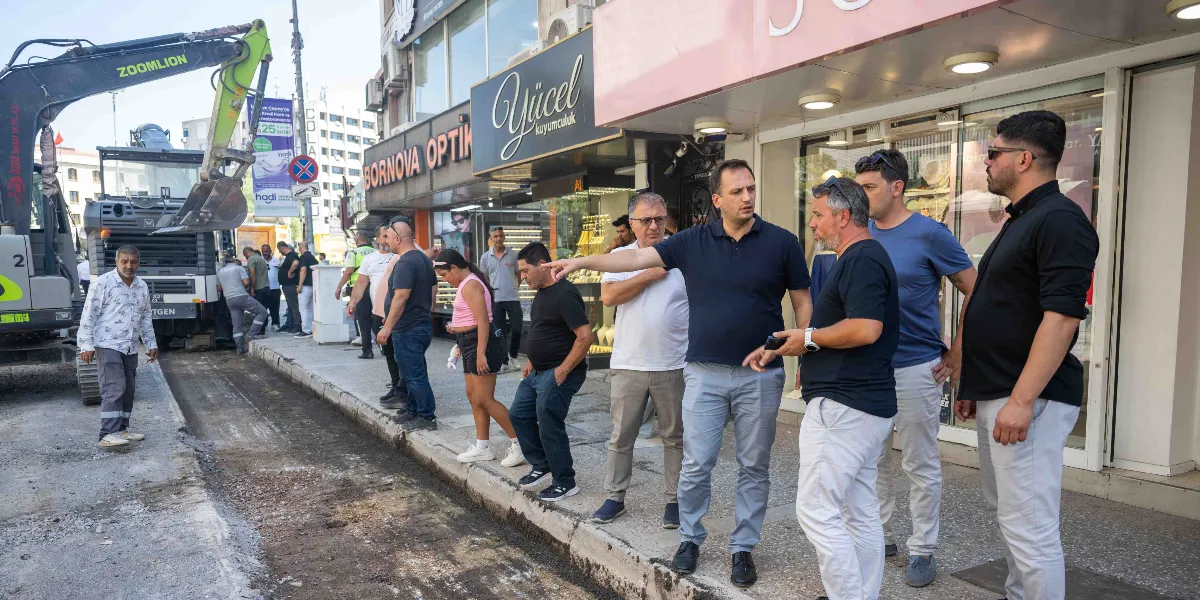 Mustafa Kemal Caddesi'ndeki yol yenileme çalışmalarını inceleyen bir grup yetkili, kaldırım üzerinde durarak inşaat alanını gözlemliyor. Grubun önünde yer alan lacivert tişörtlü bir kişi, eliyle çalışma alanını işaret ederek yanındakilere bilgi veriyor. Arka planda Bornova Optik ve Yücel Kuyumculuk tabelalarının bulunduğu dükkanlar ile yol çalışmasında kullanılan bir Zoomlion marka iş makinesi görülüyor.
