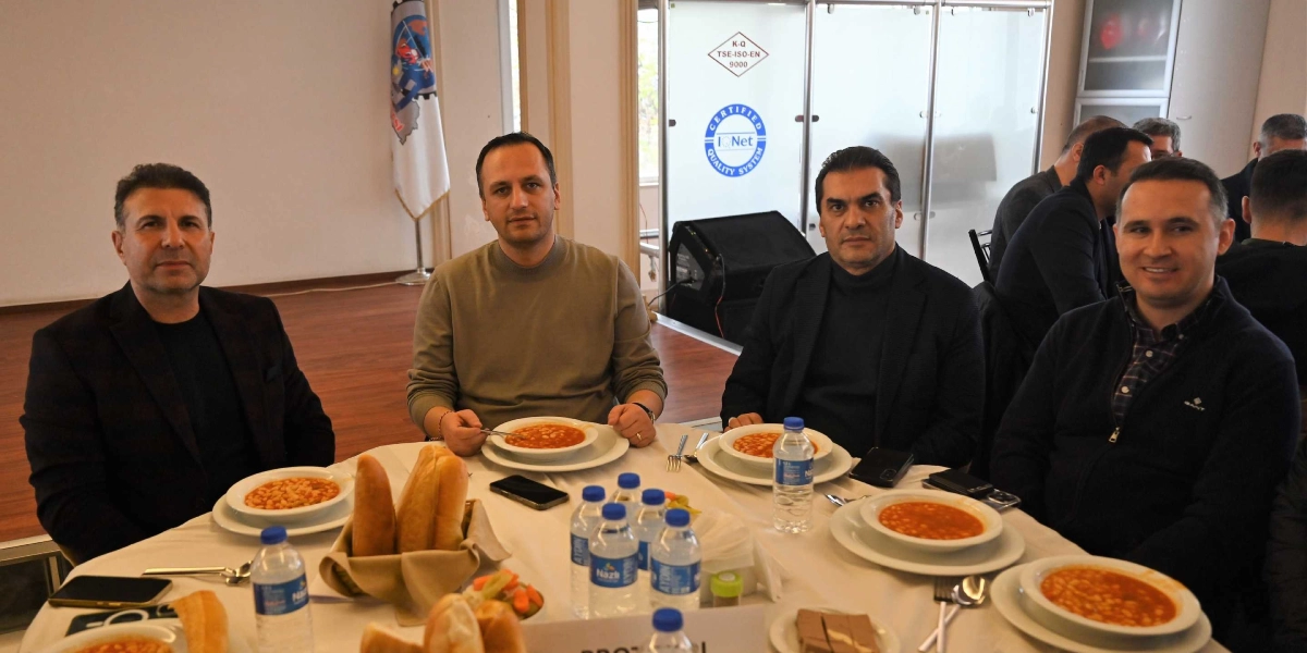 Beyaz örtülü bir masada oturan dört erkek, önlerinde kuru fasulye tabakları ve ekmeklerle yemek yiyor. Arka planda bir bayrak ve üzerinde kalite sertifikası logoları bulunan cam bölmeli bir duvar yer alıyor. Masada su şişeleri, turşu ve helva gibi ikramlar bulunurken, kişiler doğrudan kameraya bakarak poz veriyor.