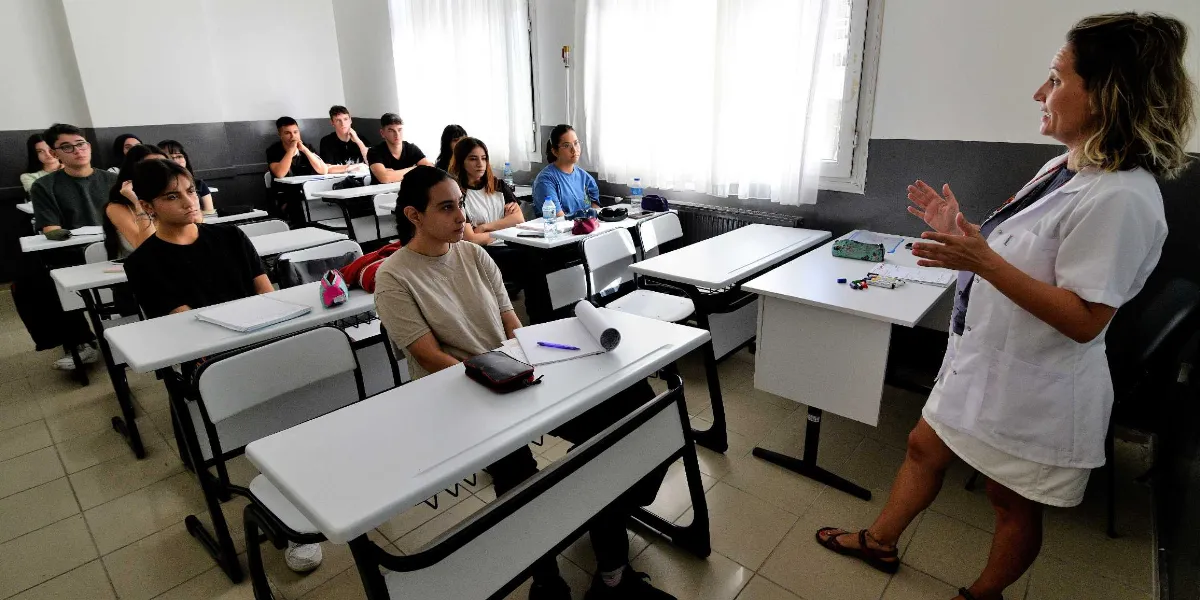 Beyaz önlüklü bir kadın eğitmen, sınıfta ders anlatan bir konumda öğrencilere hitap ediyor. Sınıfta sıralarda oturan genç öğrenciler, dikkatle eğitmeni dinliyor. Aydınlık bir sınıf ortamında, beyaz masalar ve sandalyeler düzenli bir şekilde yerleştirilmiş.