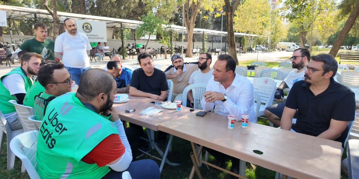 Açık havada, park benzeri bir alanda bir araya gelmiş moto kuryeler ve katılımcılar, yan yana dizilmiş masaların etrafında oturarak sohbet ediyor. Ön planda, üzerinde 