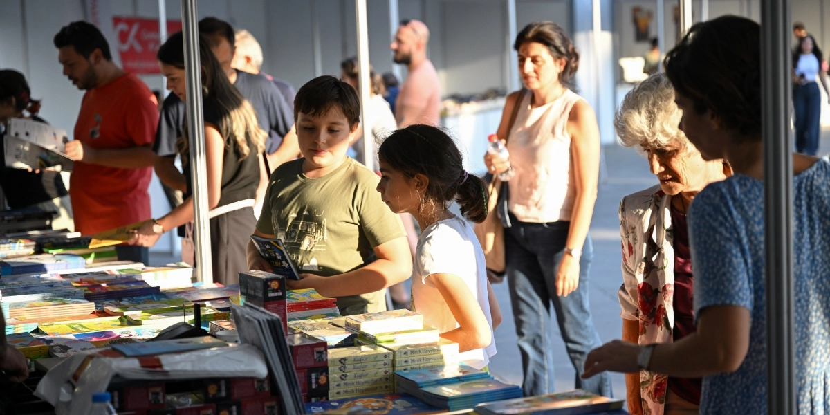 Bornova Kitap Günleri'nde stantları gezen ve kitapları inceleyen farklı yaş gruplarından ziyaretçiler. Ön planda kitaplara bakan bir erkek ve bir kız çocuğu, arka planda ise kitap stantları arasında dolaşan yetişkinler yer alıyor.