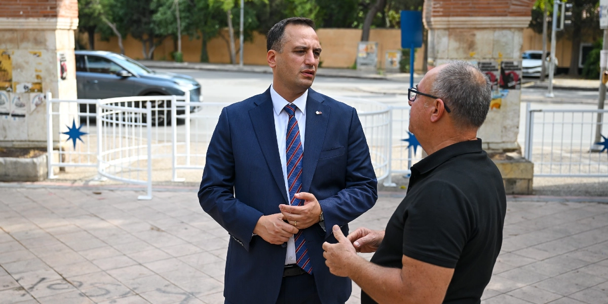 Lacivert takım elbiseli ve çizgili kravatlı bir erkek, park alanında siyah tişörtlü ve gözlüklü bir adamla ayakta durarak sohbet ediyor. Arka planda beyaz metal çitler, parkın girişindeki taş sütunlar ve park halindeki bir otomobil görülüyor.