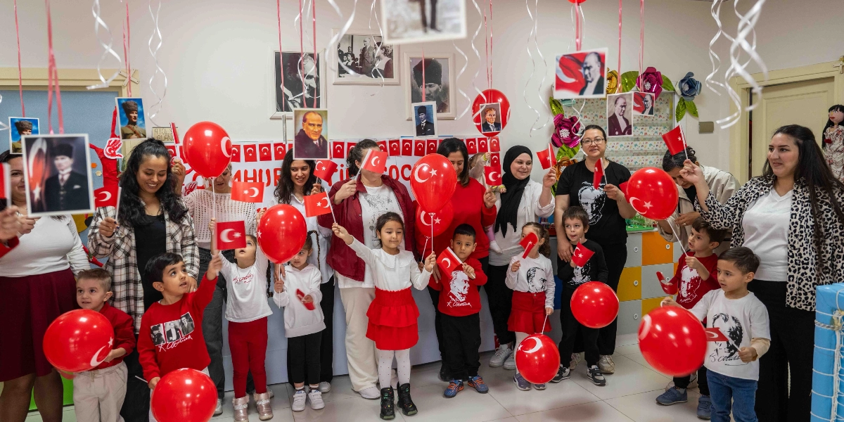 Cumhuriyet Bayramı kutlaması için bir araya gelmiş çocuklar ve yetişkinler, ellerinde Türk bayrakları ve kırmızı balonlarla poz veriyor. Çocukların bir kısmı üzerinde Atatürk baskılı tişörtler giyerken, odanın duvarlarında Atatürk fotoğrafları ve Türk bayrakları asılı. Tavandan aşağıya doğru sarkan kurdelelere bağlı küçük Atatürk fotoğrafları ve bayraklar, ortamda şenlikli bir atmosfer oluşturuyor.