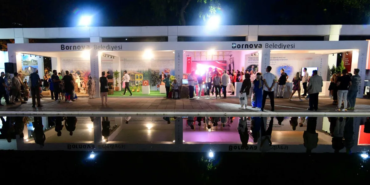 Gece vakti aydınlatılmış, üzerinde Bornova Belediyesi yazan modern ve geniş bir stant alanı. Stant, açık bölmelerden oluşuyor ve duvarlarında Bornova'nın tarihi ve kültürel değerlerini anlatan görseller ile bilgilendirme panoları yer alıyor. İçeride ve önünde çok sayıda ziyaretçi bulunuyor; bazıları panoları incelerken bazıları stant görevlileriyle sohbet ediyor. Stant alanının hemen önünde, yapının ve insanların yansımasının görüldüğü durgun bir su havuzu bulunuyor.