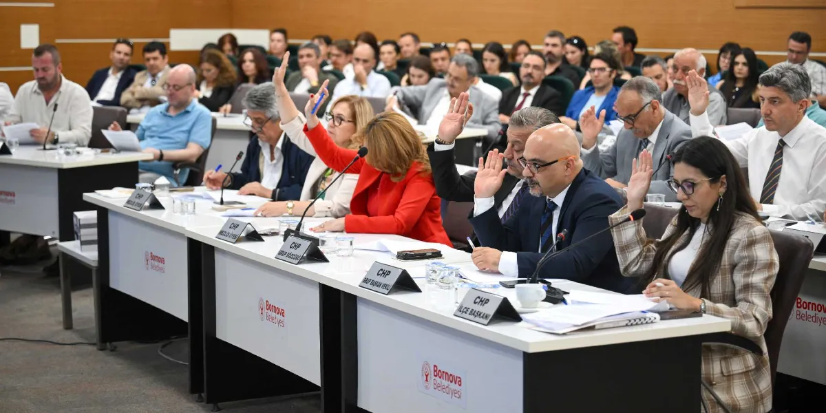 Bornova Belediyesi meclis toplantısında, masalarda oturan üyelerin büyük bir kısmının oylama yapmak için ellerini kaldırdığı görülüyor. Ön planda, üzerinde 