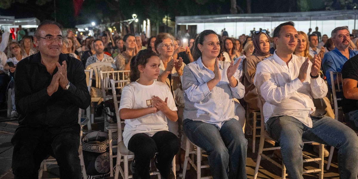 Açık havada düzenlenen bir etkinlikte, beyaz sandalyelerde yan yana oturan ve alkışlayan bir grup insan. Ön planda, siyah gömlekli bir erkek, beyaz tişörtlü bir kız çocuğu, çizgili gömlekli bir kadın ve beyaz gömlekli bir erkek yer alıyor. Arka planda ise etkinliği izleyen kalabalık bir izleyici grubu ve aydınlatılmış stantlar görülüyor.