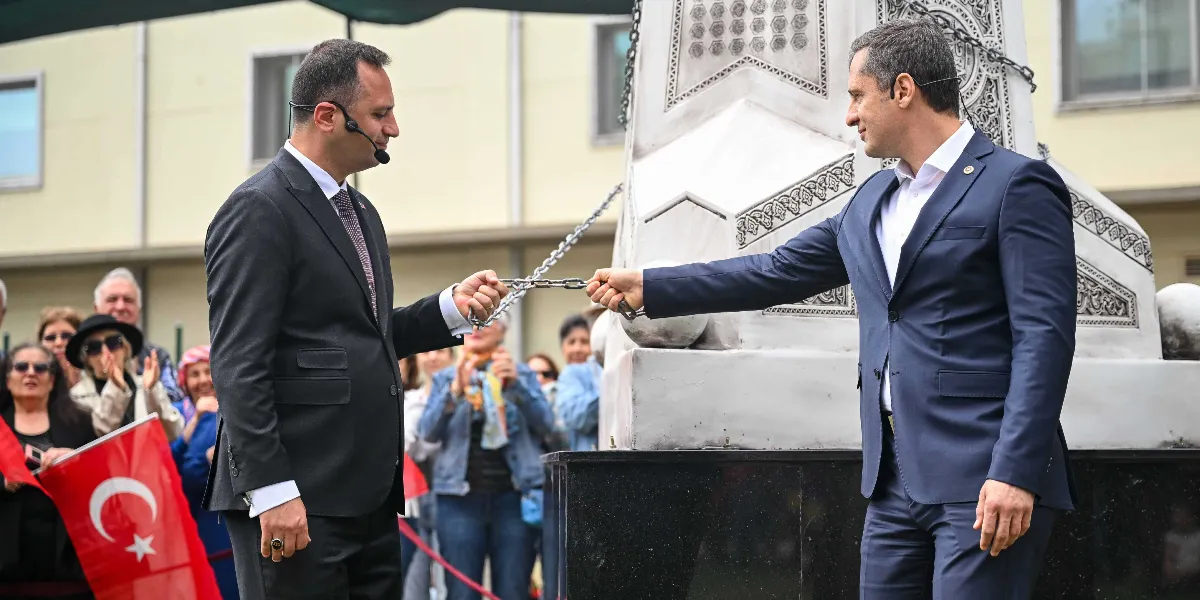 Takım elbiseli iki erkek, yanlarında bulunan anıta bağlı metal bir zinciri birlikte çekerek koparıyor. Arka planda onları izleyen ve alkışlayan bir grup insan ile ellerinde Türk bayrakları görülüyor.