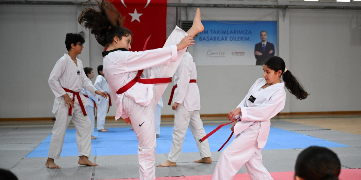 Kapalı bir spor salonunda beyaz tekvando kıyafetleri ve kırmızı kuşakları giymiş çocuklar antrenman yapıyor. Ön planda bir kız çocuğu havaya doğru yüksek bir tekme atarken, yanında başka bir kız çocuğu savunma pozisyonunda duruyor. Arka planda ise diğer öğrenciler çalışmaya devam ediyor. Duvarın bir kısmında Türk bayrağı, diğer kısmında ise üzerinde 