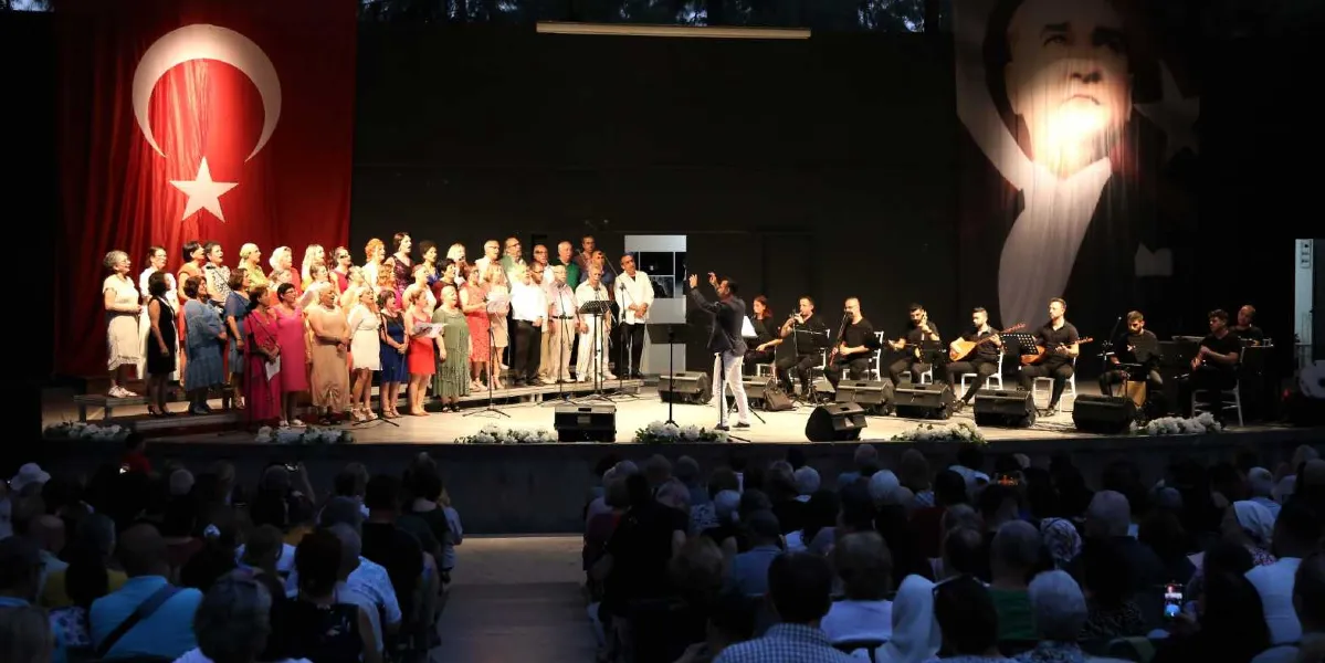 Açık hava sahnesinde, arkada büyük bir Türk bayrağı ve Mustafa Kemal Atatürk posterinin asılı olduğu bir konserde, şef yönetimindeki koro ve müzisyenler performans sergiliyor. Sahnenin sol tarafında kadınlardan oluşan kalabalık bir koro, sağ tarafında ise bağlama ve diğer enstrümanları çalan müzisyenler yer alıyor. Sahne önünde orkestra şefi bulunurken, ön planda ise konseri izleyen kalabalık bir seyirci grubu görülüyor.
