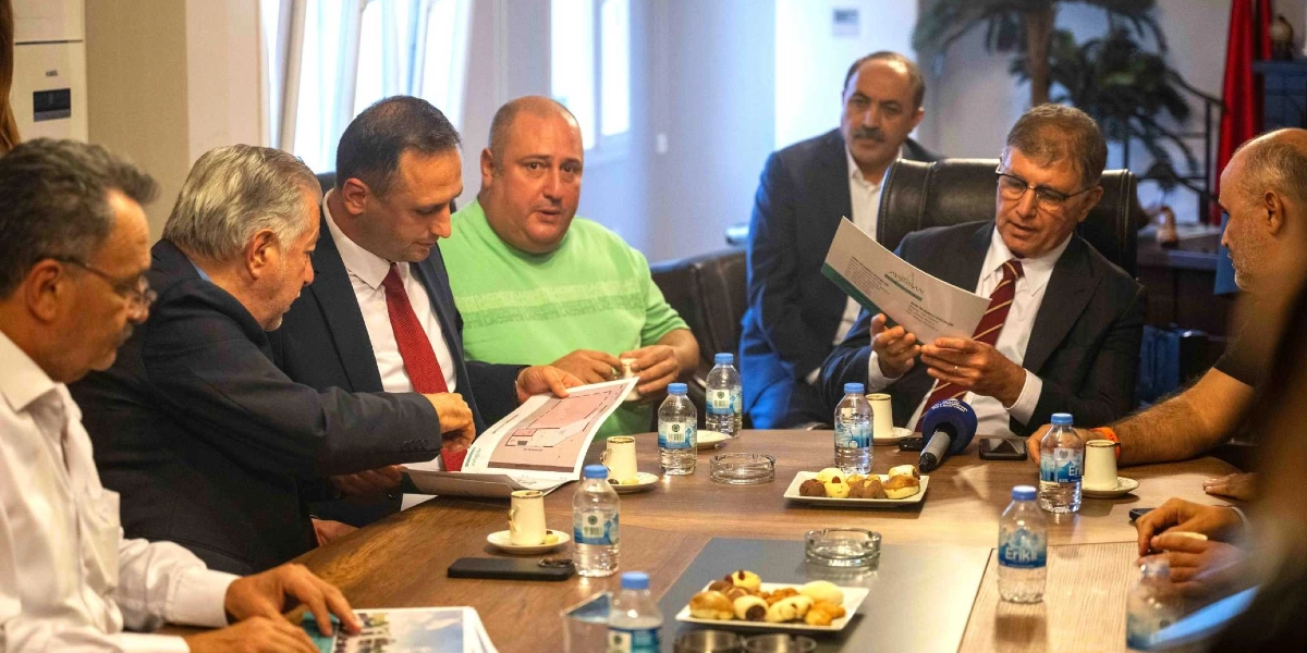 Toplantı masasında oturan bir grup erkek, önlerindeki mimari planları ve belgeleri inceleyerek fikir alışverişinde bulunuyor. Masanın üzerinde su şişeleri, Türk kahvesi fincanları ve kurabiye tabakları yer alıyor. Takım elbiseli katılımcıların yanı sıra yeşil tişörtlü bir kişi de görüşmeye eşlik ediyor.