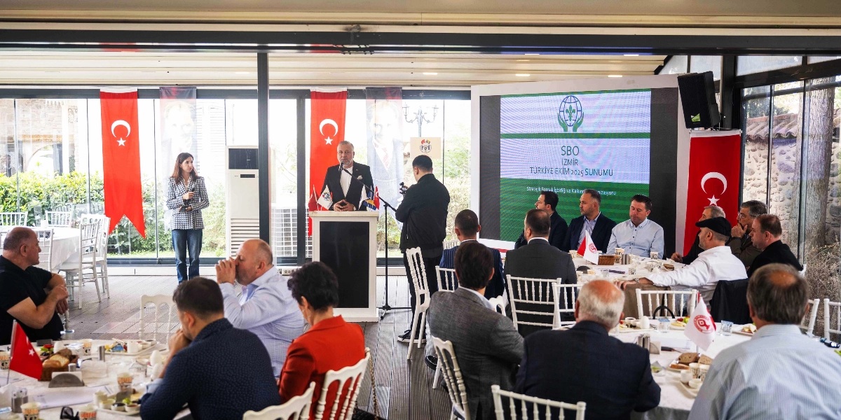 Bornova'da düzenlenen bir toplantıda, kürsüde konuşma yapan bir kişi ve onu dinleyen iş insanları yer alıyor. Salonun arka planında üzerinde 