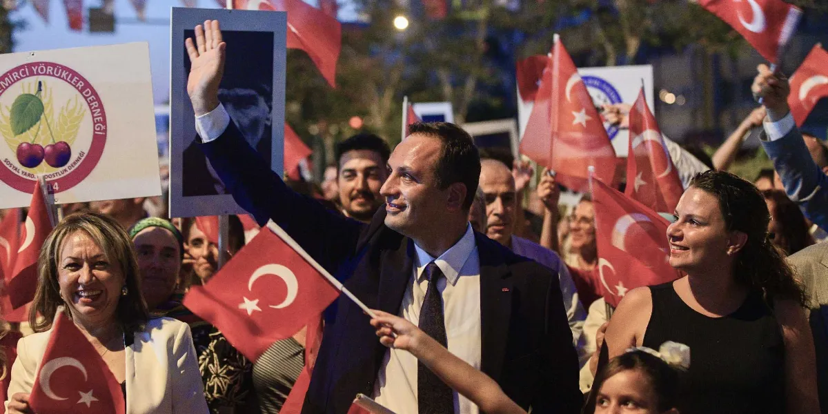 Kalabalık bir grup insan, ellerinde Türk bayrakları ve Atatürk portreleri ile bir yürüyüşte yer alıyor. Ortada, lacivert takım elbiseli ve kravatlı bir erkek el sallayarak gülümsüyor. Sol tarafta 