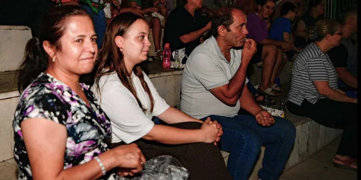 Açık havada düzenlenen bir sinema etkinliğinde, basamaklı oturma alanında yan yana oturan izleyiciler. Ön planda, çiçek desenli bluz giyen bir kadın, yanında beyaz tişörtlü genç bir kadın ve gri polo yaka tişörtlü orta yaşlı bir adam yer alıyor. İzleyiciler dikkatle karşıya bakıyor, arka planda ise etkinliği takip eden diğer insanlar seçiliyor.