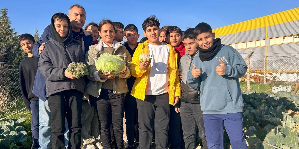Açık havada, bir sebze tarlasında yan yana duran ve ellerinde hasat ettikleri brokoli, lahana ve karnabahar gibi sebzeleri tutan bir grup öğrenci ve bir yetişkin, neşeyle kameraya gülümsüyor. Öğrencilerden biri her iki eliyle baş parmağını havaya kaldırarak poz verirken, arkada tel örgüler ve sarı renkli bir depo binası görülüyor.