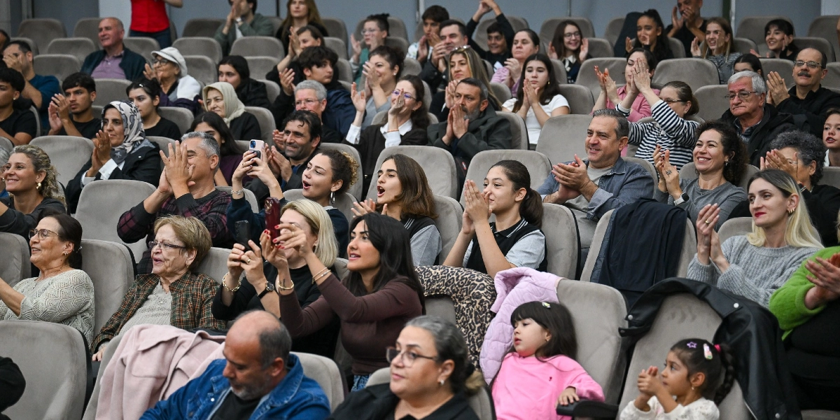 Kapalı bir salonda, gri koltuklarda oturan ve büyük bir coşkuyla alkışlayan, aralarında çocukların da bulunduğu kalabalık bir izleyici grubu. Bazı izleyiciler ellerindeki cep telefonlarıyla sahnedeki bir etkinliği kaydederken, katılımcıların çoğu gülümseyerek sahneye doğru bakıyor ve alkış tutuyor.