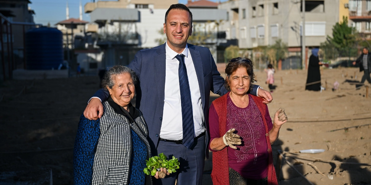 Lacivert takım elbiseli bir erkek, iki yanında duran ve ellerinde toprakla çalışmaktan kirlenmiş bahçe eldivenleri ile fide tutan iki kadınla birlikte açık havada gülümsüyor. Arka planda toprak bir alan ve yerleşim yerleri bulunuyor.