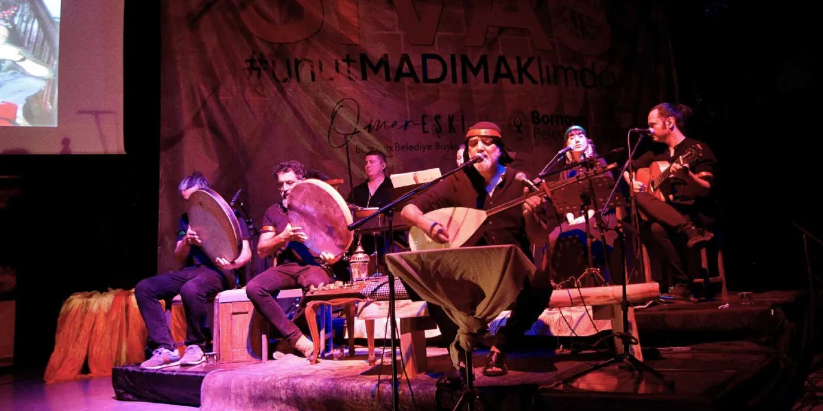 Sahne üzerinde bağlama çalan bir sanatçı ve ona eşlik eden müzisyenlerden oluşan bir grup, loş ve kırmızı ışıklarla aydınlatılmış bir ortamda performans sergiliyor. Müzisyenler arasında el davulu çalanlar ve gitarist bulunuyor. Arka planda üzerinde 