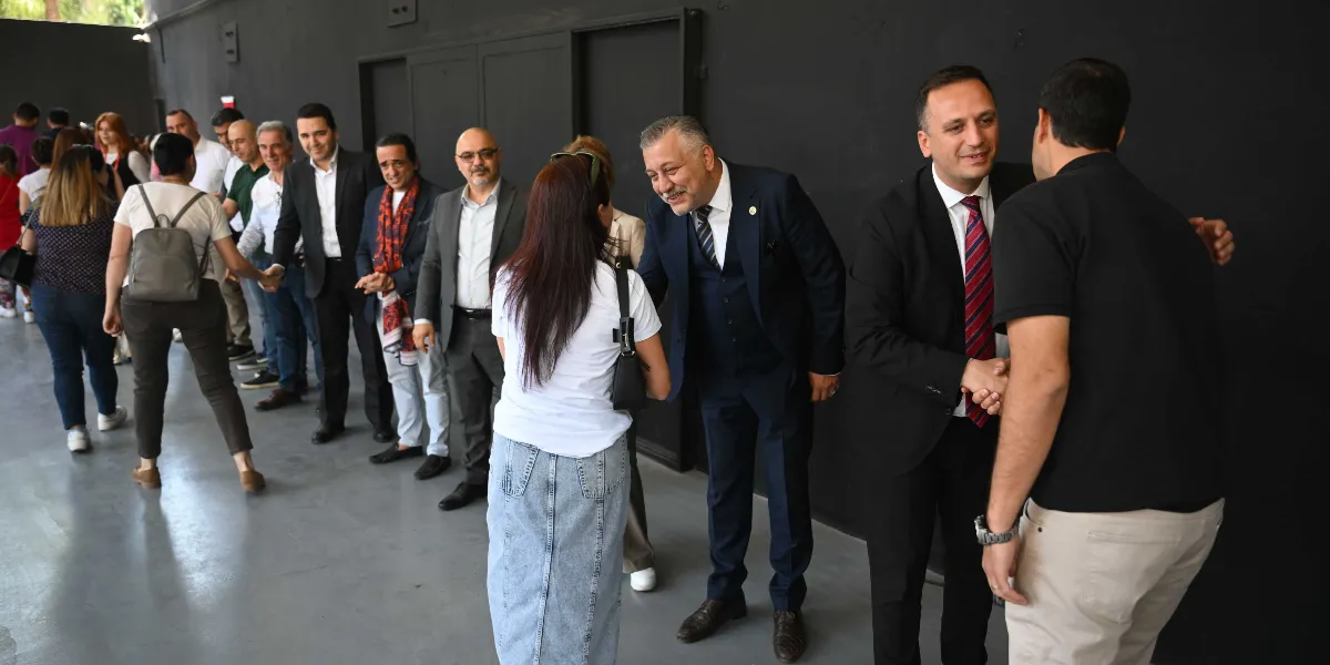 Siyah bir duvarın önünde, resmi kıyafetli bir grup insanın birbiriyle bayramlaştığı bir etkinlik. Ön planda takım elbiseli iki kişi, gelen konuklarla tokalaşarak onları karşılıyor. Arka planda ise sıraya girmiş veya birbiriyle sohbet eden kadın ve erkeklerden oluşan bir kalabalık görülüyor.