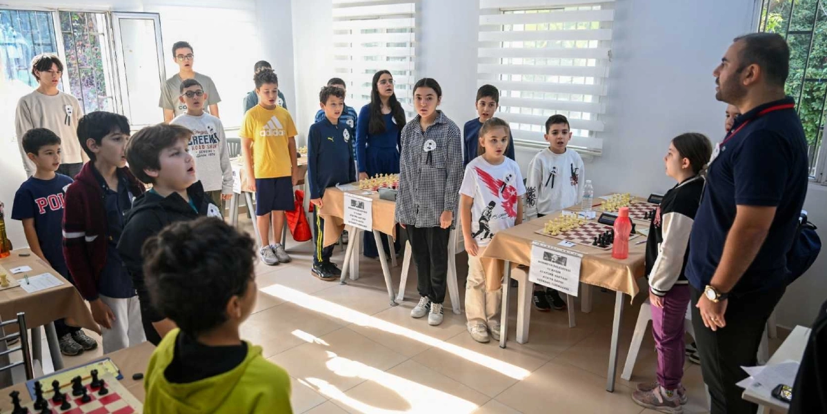 Aydınlık bir sınıfta, Bornova Belediyesi tarafından düzenlenen 10 Kasım Atatürk Haftası Satranç Turnuvası kapsamında bir araya gelmiş çocuklar ve gençler, saygı duruşunda bulunuyor. Odadaki satranç masalarının üzerinde üzerinde etkinlik bilgilerinin yer aldığı kağıtlar asılıyken, katılımcılar ciddiyetle ayakta duruyor. Sağ tarafta bir eğitmen veya görevli, öğrencilere dönük şekilde ayakta bekliyor.