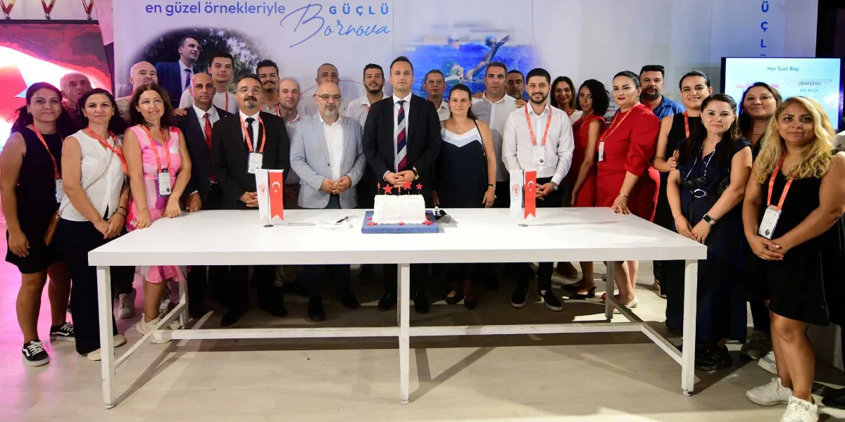 Uluslararası İzmir Fuarı'ndaki Bornova Belediyesi standında, üzerinde kırmızı yıldız süslemeli beyaz bir pasta bulunan uzun bir masanın arkasında yan yana duran, resmi kıyafetli ve yaka kartlı belediye çalışanları ve yetkililerden oluşan kalabalık bir grup. Arka planda 